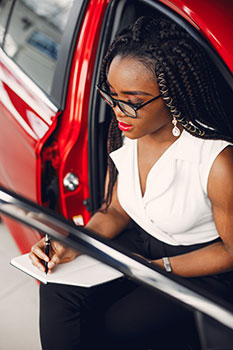 Woman sitting in a red car writing down her  budget for car maintenance and auto insurance in a notebook